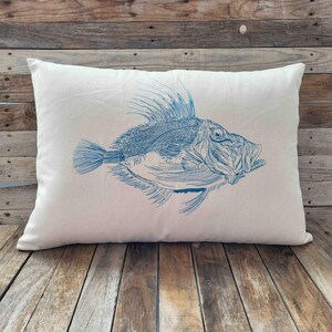 Könnte beinhalten: Ein rechteckiges weißes Kissen mit einer detaillierten blauen Illustration eines Fisches. Der Fisch hat stachelige Flossen und einen offenen Mund, wobei filigrane Linien eine strukturierte Optik erzeugen. Das Kissen liegt auf einer Holzoberfläche.