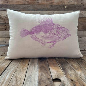 Könnte beinhalten: Ein rechteckiges Kissen mit cremefarbenem Hintergrund. Eine detaillierte lila Illustration eines Fisches ist auf das Kissen gedruckt. Der Fisch hat markante Flossen und einen detaillierten Körper. Das Kissen liegt auf einer Holzoberfläche.