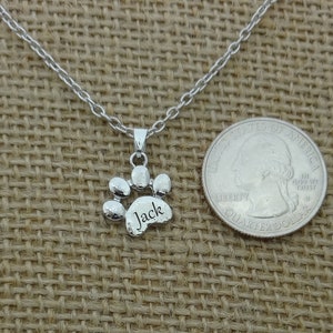 May include: A silver-colored paw print pendant necklace with the name "Jack" engraved on the paw. The necklace is displayed on a tan burlap surface next to a U.S. quarter for size comparison.