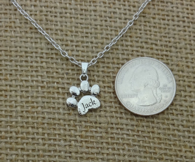 May include: A silver-colored paw print pendant necklace with the name "Jack" engraved on the paw. The necklace is displayed on a tan burlap surface next to a U.S. quarter for size comparison.
