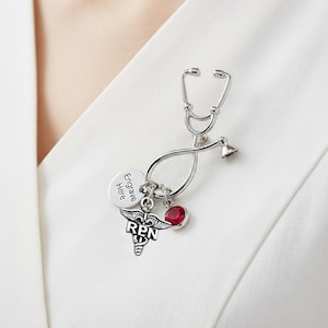 May include: A silver stethoscope brooch with a red gemstone and a Registered Practical Nurse (RPN) emblem. The brooch includes a charm that says "Engrave Here". This medical-themed accessory is designed to be worn on clothing.