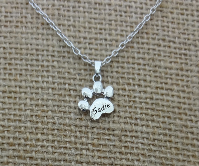 May include: A silver-colored necklace featuring a paw print pendant. The pendant has the name "Sadie" engraved on it. The necklace is displayed on a neutral-colored fabric background.