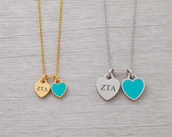 May include: Two necklaces, one gold-toned and one silver-toned, each featuring two heart-shaped charms. One charm on each necklace is engraved with the letters ZTA, while the other charm is filled with a bright turquoise enamel color.