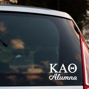 May include: White vinyl decal with the Greek letters KAO and the word "Alumna" in a cursive font. The decal is affixed to the rear window of a vehicle, with a reflection of clouds visible.