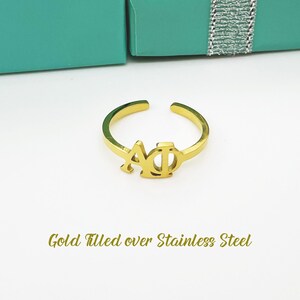 Könnte beinhalten: Verstellbarer goldfarbener Ring mit den griechischen Buchstaben ΑΦ. Der Ring besteht aus goldgefülltem Material über Edelstahl. Der Ring wird auf einer weißen Oberfläche mit einer türkisfarbenen Box im Hintergrund präsentiert.