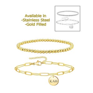 May include: Two gold-coloured bracelets are shown on a white background. One is a beaded style, and the other features a chain link design with a round charm that reads "KAΘ". The text "Available in -Stainless Steel -Gold Filled" is also visible.