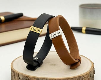 May include: Two leather bracelets, one black and one brown, each featuring a rectangular metal charm engraved with the Greek letters Pi Kappa Phi. The black bracelet has a gold-tone charm, while the brown bracelet has a silver-tone charm.