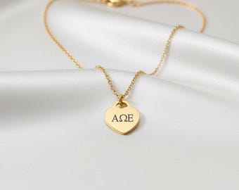 May include: A gold necklace featuring a heart-shaped pendant with the letters AΩE. The necklace is presented on a white, silky fabric. The chain and pendant are gold in colour.