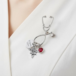 May include: Silver stethoscope pin with a red gemstone and a charm with the text "Engrave Here". The pin also features a medical symbol with the letters "ADN".