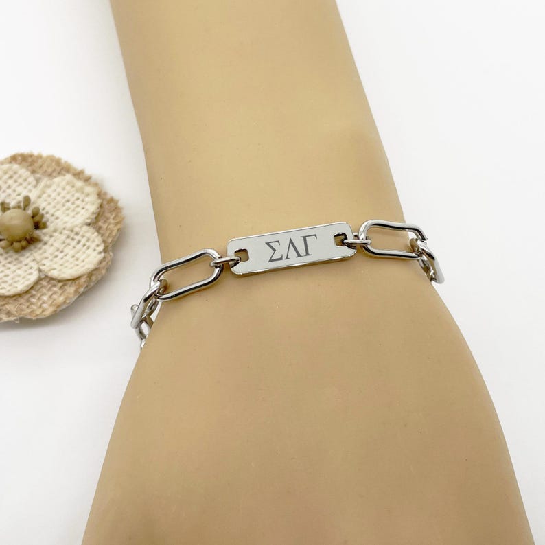 Peut inclure: Un bracelet cha&icirc;ne argent&eacute; avec une plaque rectangulaire grav&eacute;e des lettres "&Sigma;&Lambda;&Gamma;". Le bracelet est compos&eacute; de maillons m&eacute;talliques rectangulaires. Le bracelet est port&eacute; au poignet.