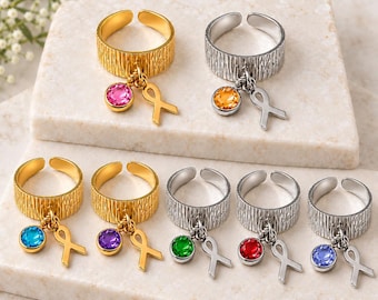 May include: A collection of adjustable rings featuring a textured band, each adorned with a small dangling awareness ribbon charm and a single round faceted gemstone in various colors including pink, orange, blue, purple, green, red, and light blue.