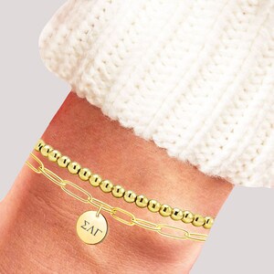May include: Gold chain bracelet with a round charm that says "&Sigma;&Lambda;&Gamma;". The bracelet is layered with a gold beaded bracelet.