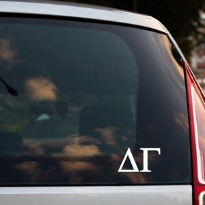 May include: A white vinyl decal with the Greek letters Delta Gamma on a silver car window.