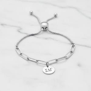 Puede incluir: Pulsera de cadena plateada con un dije redondo. La pulsera presenta eslabones ovalados alargados y una cadena más fina. El dije tiene letras griegas grabadas. La pulsera tiene un cierre deslizante ajustable. Metal brillante.