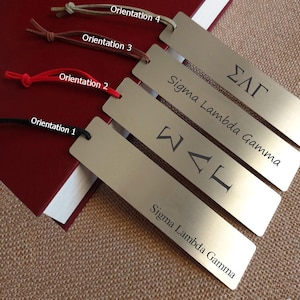 May include: Four metal bookmarks with different colored cords. Each bookmark has the Greek letters ΣΑΓ or ΣΛΓ and the words "Sigma Lambda Gamma" engraved on it. The bookmarks are resting on a red book.