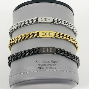 May include: Three stainless steel chain bracelets in silver, gold, and black. Each bracelet features a rectangular plate with Greek letters. The image also includes text that reads "Stainless Steel *Hypoallergenic *Non-Tarnishing."