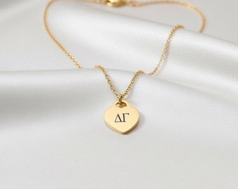 May include: A gold necklace featuring a heart-shaped pendant with the Greek letters ΔΓ. The fine chain and clasp are visible. The pendant hangs on a white, silky fabric. The necklace is a delicate piece of jewellery.