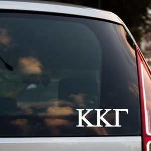 May include: A white vinyl decal with the letters "KKΓ" on a silver car window.