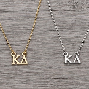 May include: Two necklaces featuring the Greek letters Kappa and Delta as pendants. One necklace is gold-toned with a matching chain, and the other is silver-toned with a matching chain. Both pendants are suspended from delicate link chains.