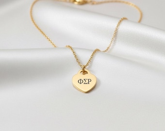 May include: A gold necklace featuring a heart-shaped pendant with the Greek letters ΦΣΡ. The necklace is presented on a white, satin-like material.