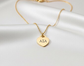 May include: A gold-coloured necklace featuring a heart-shaped pendant. The pendant is engraved with the letters "ASA". The necklace is presented on a white, silky fabric.
