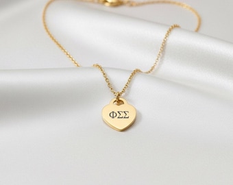May include: A gold necklace featuring a heart-shaped pendant with the Greek letters ΦΣΣ in black. The fine chain and pendant are set against a backdrop of white, silky fabric. The necklace is a delicate piece of jewellery.