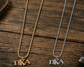May include: Two necklaces featuring the Greek letters Pi Kappa Alpha, abbreviated as ΠΚΑ. One necklace is gold-toned with a box chain, and the other is silver-toned with a curb chain. Both pendants are metal and hang from their respective chains.
