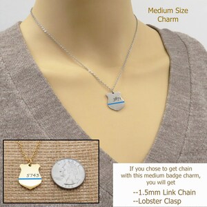 May include: A silver necklace with a silver shield charm featuring a blue line and the number "3871". A smaller gold shield charm with a blue line and the number "5743" is shown next to a UK 20 pence coin for size comparison. The necklace is 1.5mm link chain with a lobster clasp.