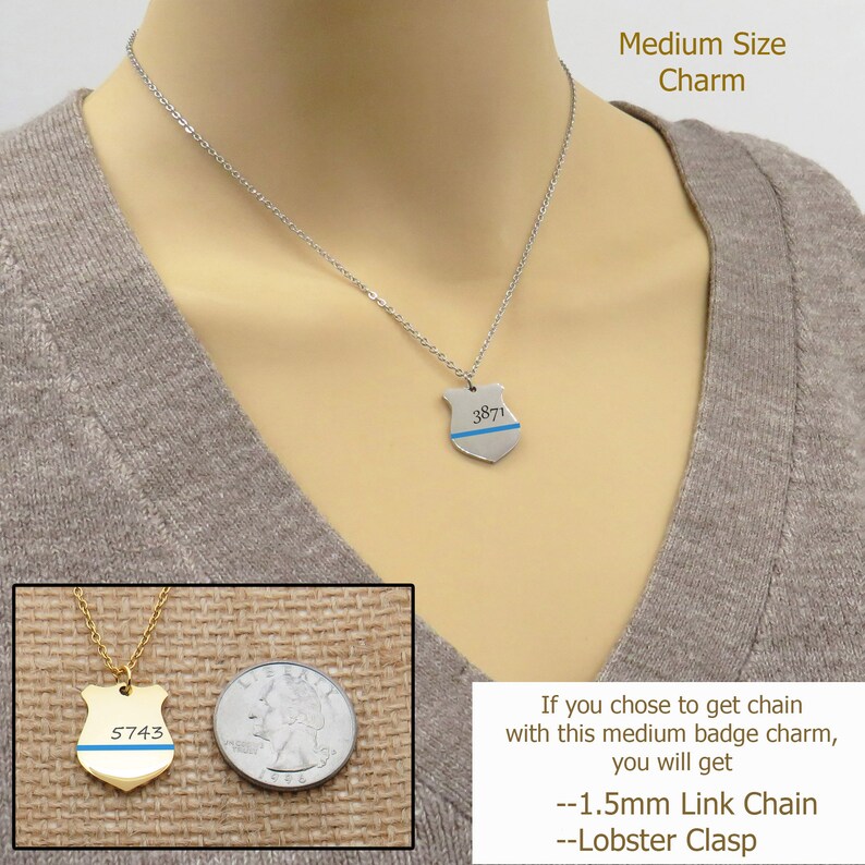 May include: A silver necklace with a silver shield charm featuring a blue line and the number "3871". A smaller gold shield charm with a blue line and the number "5743" is shown next to a UK 20 pence coin for size comparison. The necklace is 1.5mm link chain with a lobster clasp.