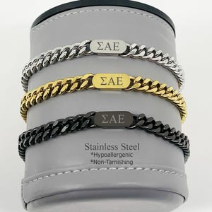 May include: Three stainless steel chain bracelets in silver, gold, and black. Each bracelet has a rectangular plate with the Greek letters ΣAE. The text "Stainless Steel *Hypoallergenic *Non-Tarnishing" is also visible.
