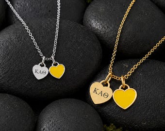 May include: Two necklaces, one silver-toned and one gold-toned, each featuring two heart-shaped charms. One charm on each necklace is engraved with the Greek letters KAΘ, while the other charm is filled with a solid yellow enamel color.