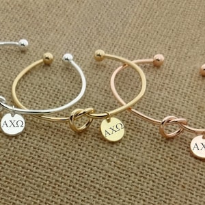 May include: Three gold knot bangle bracelets with a charm that says "AXO" on each. The bracelets are silver, gold, and rose gold.