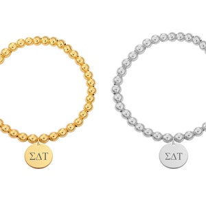 Puede incluir: Dos pulseras de cuentas con un dije. Una pulsera es dorada y la otra plateada. Ambos dijes son redondos y tienen las letras "ΣΔΤ" grabadas.