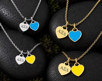May include: Four necklaces featuring two heart-shaped charms each. Two necklaces are silver-toned and two are gold-toned. Each set includes one heart engraved with the Greek letters ΑΞΔ and one solid heart in either bright blue or yellow enamel.