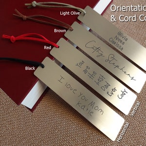 May include: Five silver bookmarks with different engraved messages. The bookmarks are arranged vertically with the top bookmark at the top of the image. The bookmarks are attached to cords of different colours: black, red, brown, and light olive. The text on the bookmarks reads: "Olivia Nora Clarissa", "Cathy Stephens", "You are the best Dad Ever", "I love you Mom Katie", and "Horizontal Vertical".