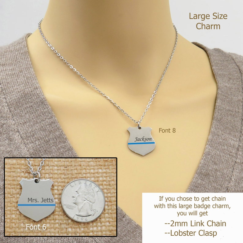 May include: A silver necklace with a large, shield-shaped charm. The charm has a blue line across it and the name "Jackson" engraved on it. The necklace is shown on a model wearing a brown sweater. The text "Font 8" is visible below the charm. The image also shows a smaller version of the charm with the name "Mrs. Jetts" engraved on it. The text "Font 6" is visible below the charm. The text "Large Size Charm" is visible in the top right corner of the image. The text "If you chose to get chain with this large badge charm, you will get --2mm Link Chain --Lobster Clasp" is visible in the bottom right corner of the image.