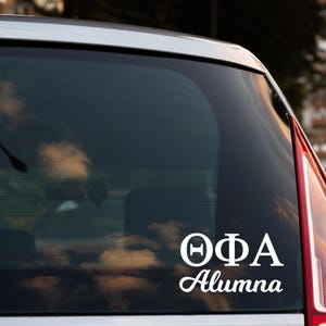 May include: White vinyl decal applied to a car window. The decal features the Greek letters Theta, Phi, and Alpha, with the word "Alumna" in a script font below. The car window reflects a cloudy sky.