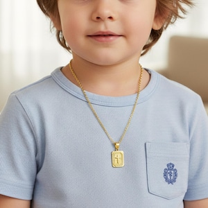 May include: A gold-colored rectangular pendant necklace with a cross design is worn over a light blue t-shirt. The pendant hangs from a delicate chain. The t-shirt has a pocket with a crest design.