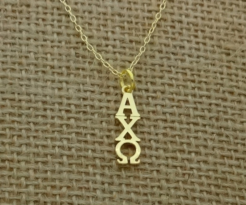 May include: A gold-colored necklace with a pendant featuring the Greek letters Alpha, Chi, and Omega. The pendant is suspended from a delicate gold chain. The background is a textured, tan fabric.