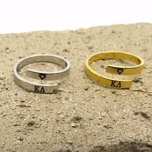 May include: Two wrap rings, one silver and one gold, are displayed on a textured surface. Each ring features a heart symbol and the letters KΔ. The rings have an open design, with the band overlapping.