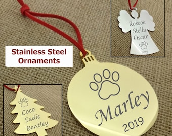 May include: Three gold stainless steel Christmas ornaments with engraved paw prints and names. The ornaments are shaped like a tree, a bauble, and an angel. The text on the ornaments reads "Coco Sadie Bentley", "Marley 2019", and "Roscoe Stella Oscar 2019".