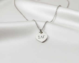 May include: A silver necklace featuring a heart-shaped pendant with the Greek letters ΣΛΓ. The necklace is presented on a white, silky fabric, with the chain and clasp visible.
