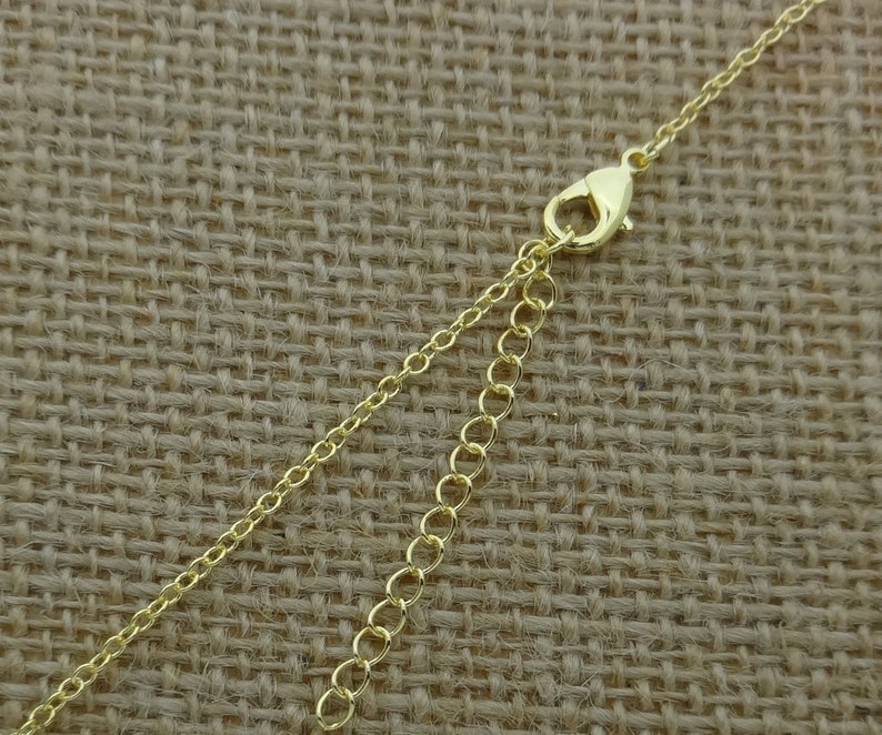 May include: A gold-colored necklace chain with a lobster clasp and an extender chain. The chain is made of small, delicate links. The background is a textured, tan fabric.