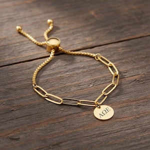 Peut inclure: Bracelet doré avec un design à maillons de chaîne et un charm circulaire. Le charm porte les lettres grecques ΑΩΕ. Le bracelet est doté d'un fermoir coulissant réglable. Le bijou est présenté sur une surface en bois.