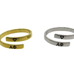 Könnte beinhalten: Zwei verstellbare Ringe, einer in Gold und einer in Silber, mit einem Herz und den eingravierten Buchstaben "AΦ".
