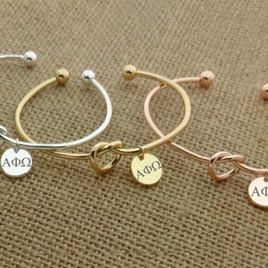 May include: Three gold knot bangle bracelets with a round charm that says "AΦΩ". The bracelets are silver, gold, and rose gold.