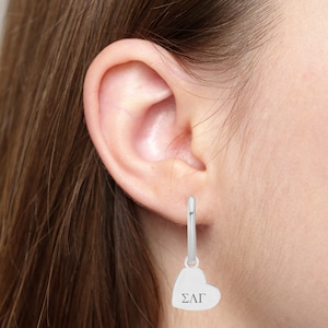 May include: Silver hoop earrings with a heart-shaped charm that says "ΣΛΓ" hanging from it.