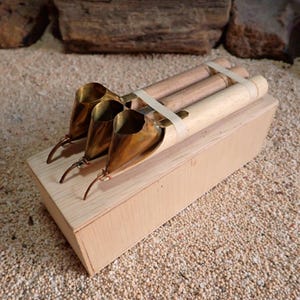 Peut inclure: Trois outils tjanting avec des réservoirs en laiton et des manches en bois, fixés ensemble par un élastique blanc, posés sur un bloc de bois rectangulaire clair. Chaque outil présente un petit bec courbé pour appliquer la cire en batik.