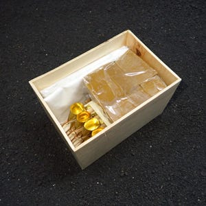 Peut inclure: Une boîte en bois contenant plusieurs articles. À l'intérieur, il y a plusieurs objets dorés avec un dessus bulbeux, un paquet de carrés de couleur ambre et un tissu blanc. La boîte est en bois clair.