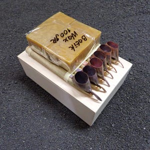 May include: A rectangular block of yellow batik wax weighing 100g in clear plastic packaging, placed on a light-coloured wooden box alongside five copper tjanting tools with bamboo handles, arranged in a row for textile art applications.